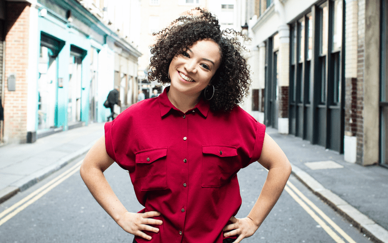 Lynette Linton standing in the middle of a street, with a bright raspberry red top on, hands on hips, smiling at the camera with their head tilted.