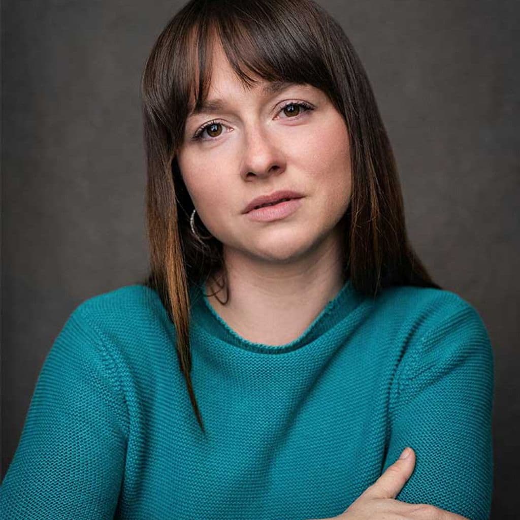 Headshot of Rosie Sheehy – a white woman in a teal shirt with her arms crossed. Her dark brown hair frames her face, with curtain fringe on her forehead.