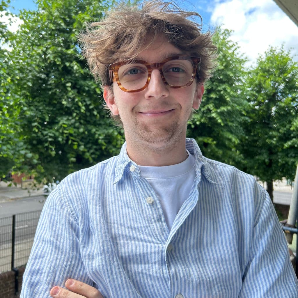 Charles, a white male with tousled hair wearing a blue and white striped shirt, stands outdoors with arms crossed, surrounded by green trees and blue skies.