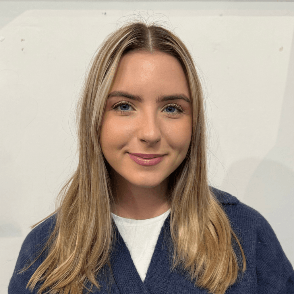 A white woman with blonde hair, blue eyes, smiling with her moth closed. Wearing a white t-shirt with a dark blue v-neck jumper.
