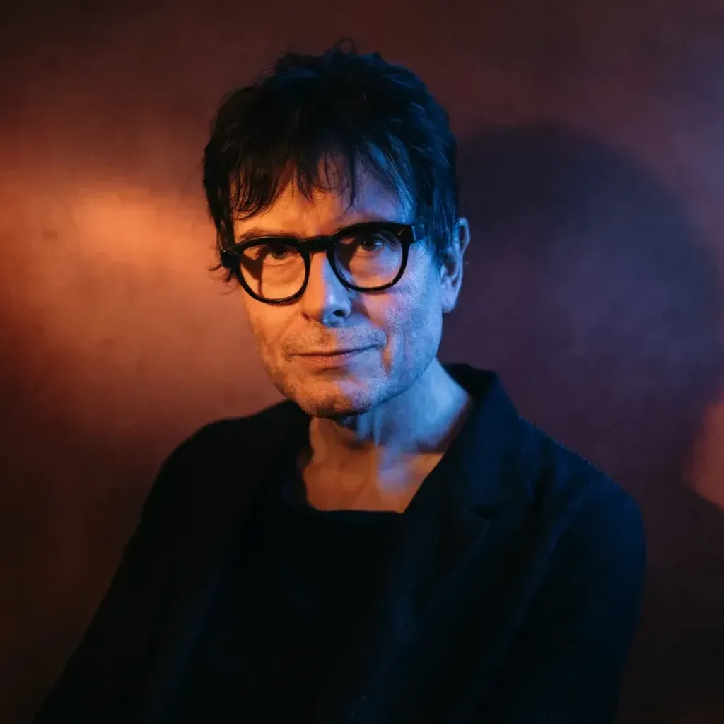 Director, James Macdonald in black rimmed glasses and dark clothes, standing against a background which is lit to shimmer gold and purple. Light shining on to James' face giving a soft glow.