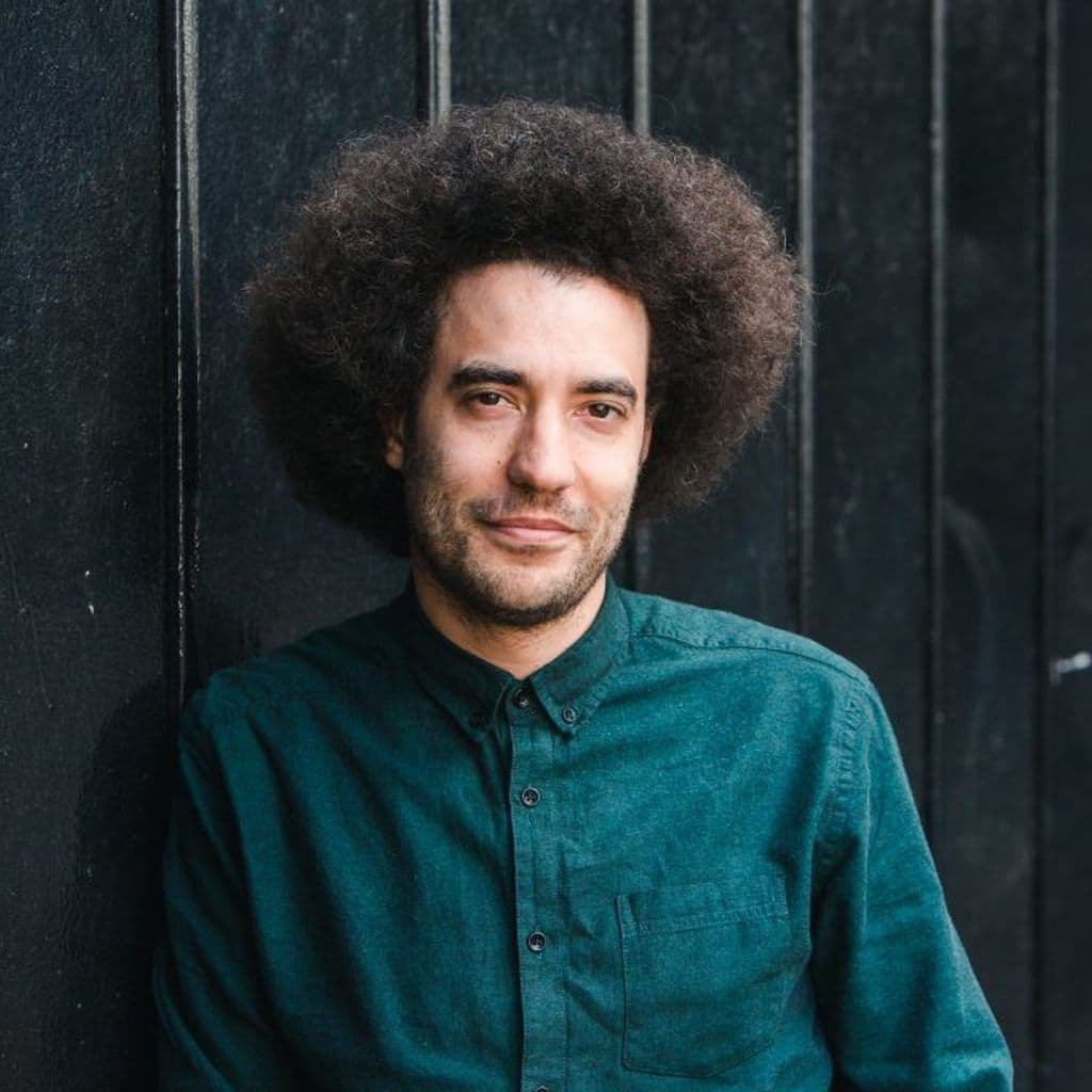 Justin Audibert's headshot, stood in front of black grates with a dark green shirt on.