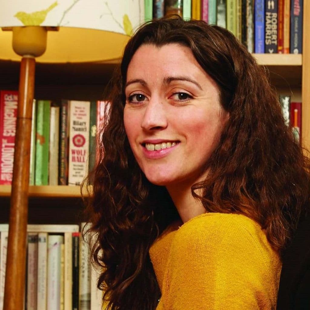 Headshot of Kate McGrath - Producer of Fuel, White woman with brown hair looking over shoulder into camera. Wearing a mustard yellow top. In front of a set of books.