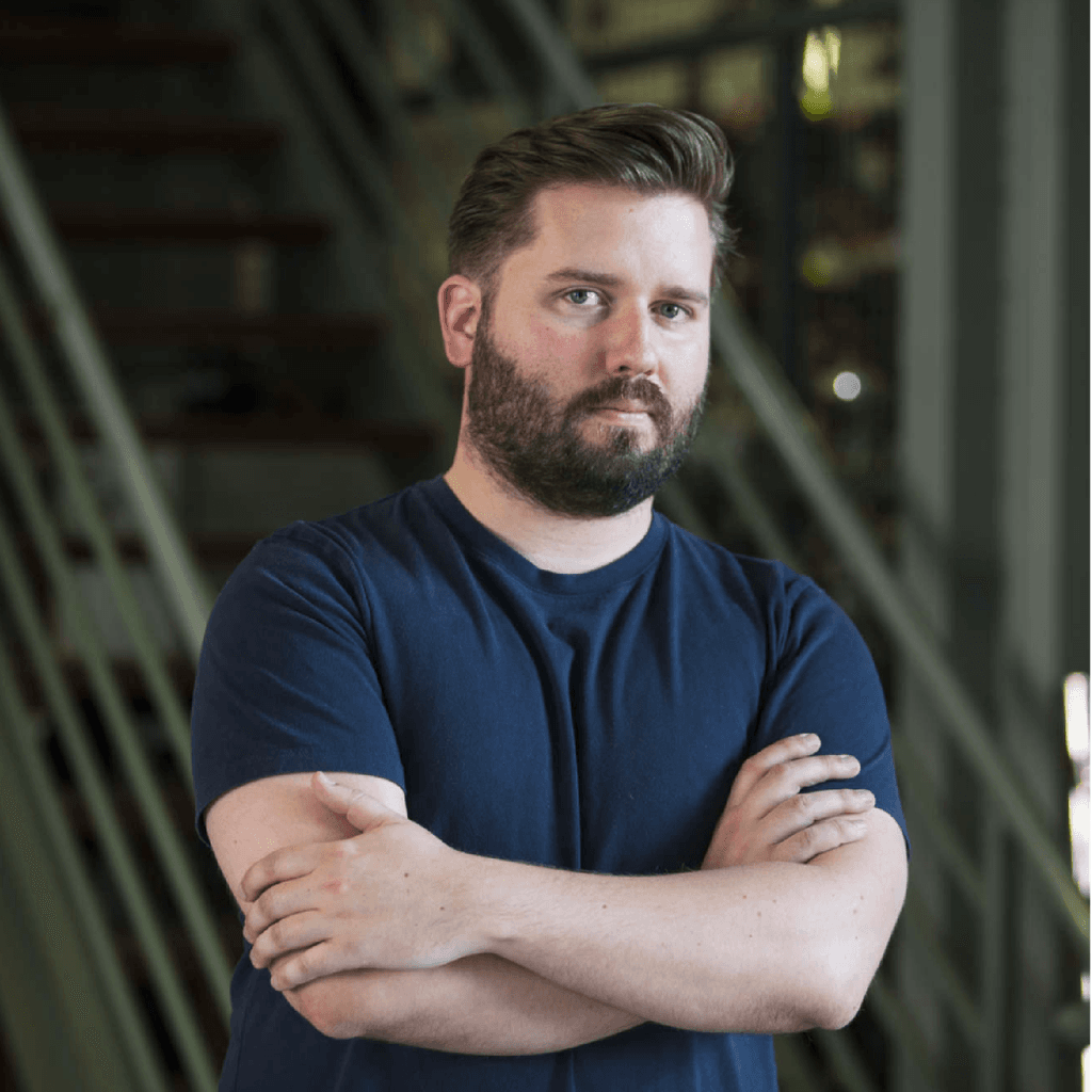 Director, Kip Williams, stood with his arms crossed looking into the camera in front of a set of stairs. Kip is a white man with dark brown hair and a full beard. He is wearing a dark blue T-shirt.