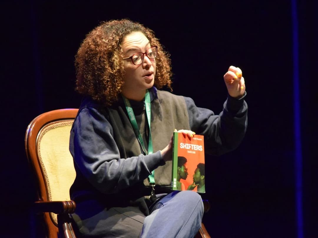Lynette Linton seated on a chair holding a script titled "Shifters" and gestures while discussing it during a masterclass.