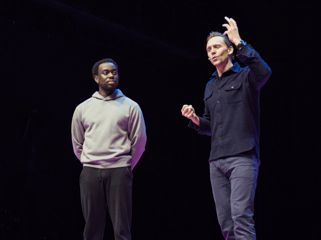 Tom Hiddleston and Richard Adetunji stand on a stage, Tom animatedly speaking while Richard listens intently, with dramatic lighting in the background.