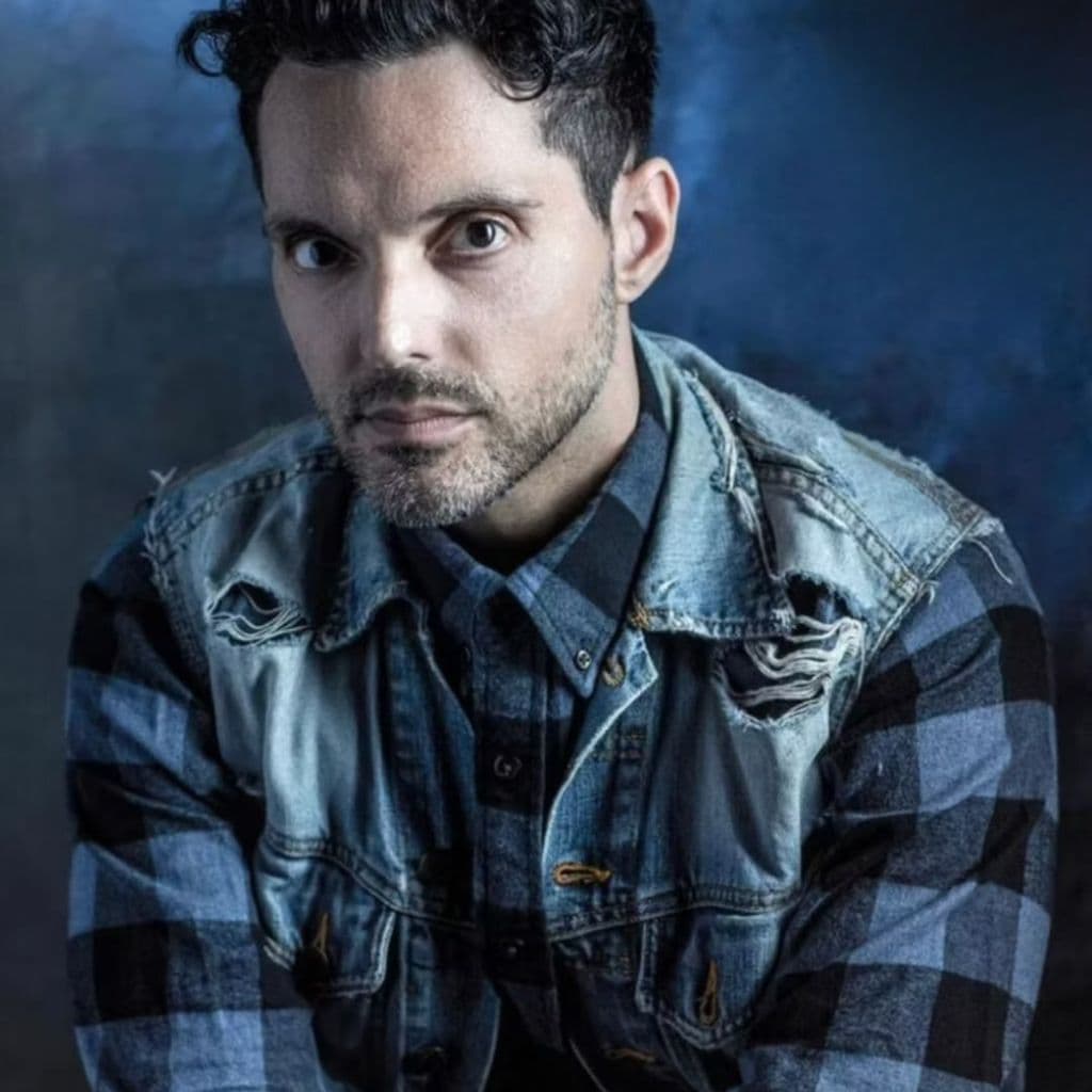 A man with short dark hair and a beard stares intently at the camera. He wears a plaid shirt under a distressed denim jacket, with a moody, blue-toned background.
