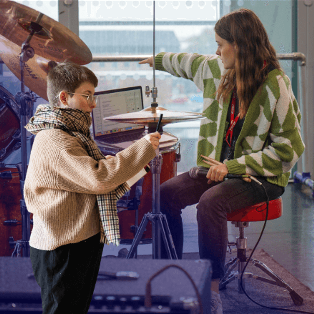 Theatremaker Jodie Irvine playing the drums and Dramaturg, writer & Director Clodagh Chapman cut out of her directing. Set in a room with windows.