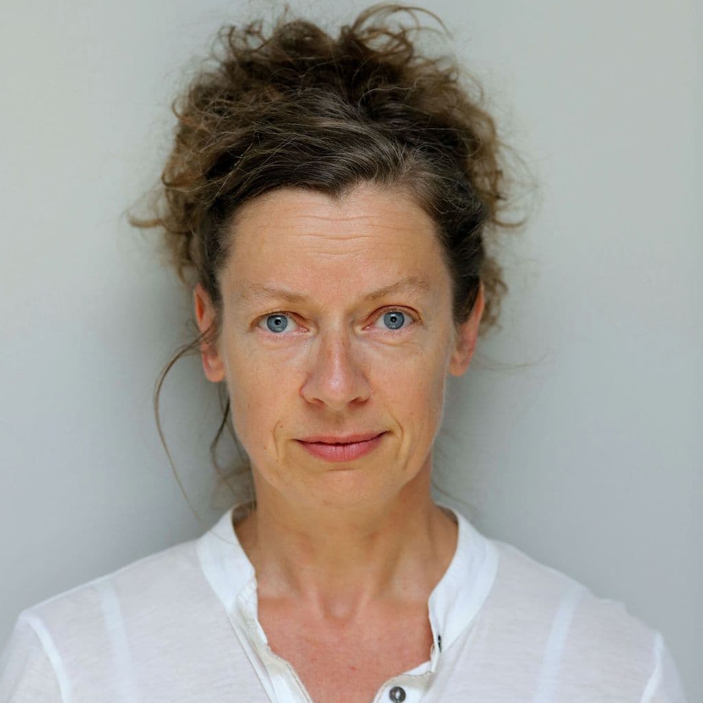 Sinéad Rushe Headshot - White woman with blue eyes and brown hair wearing a white top against a pale background.