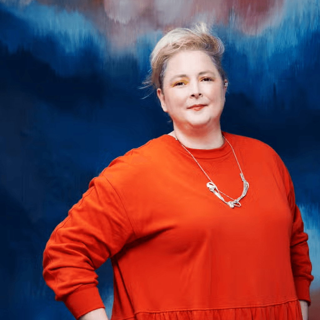Siobhan McSweeney wearing a orange jumper against a blue/purple background