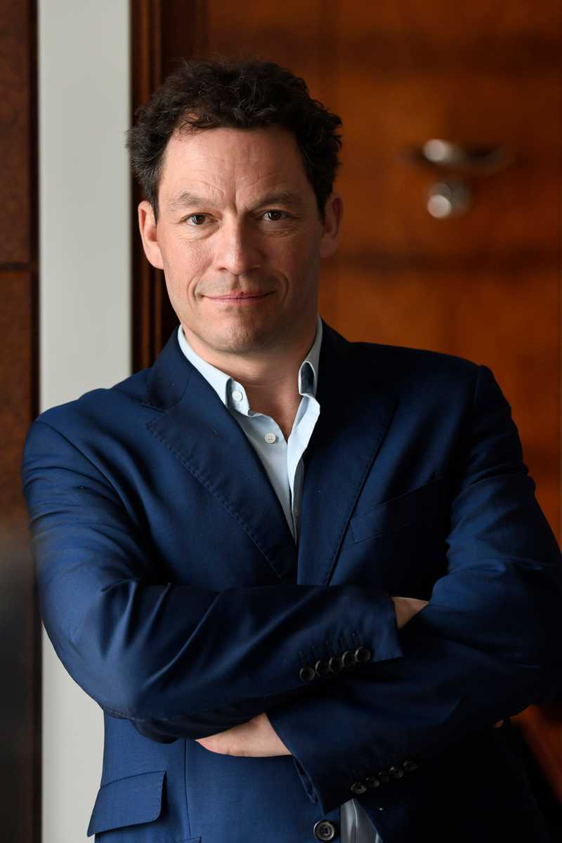 A headshout of Dominic West in a dark blue suit and white shirt, looking directly in to the camera. A blurred background of dark wood.