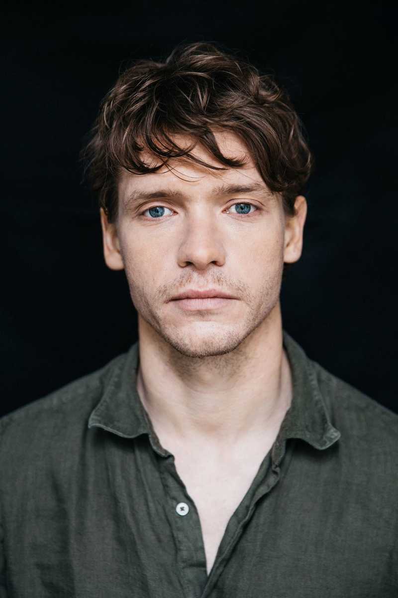 Headshot of Billy Howle, a white actor with dark red hair, blue eyes and slight stubble. Wearing a grey/green shirt open to the second button. Shot against a black bacground. 