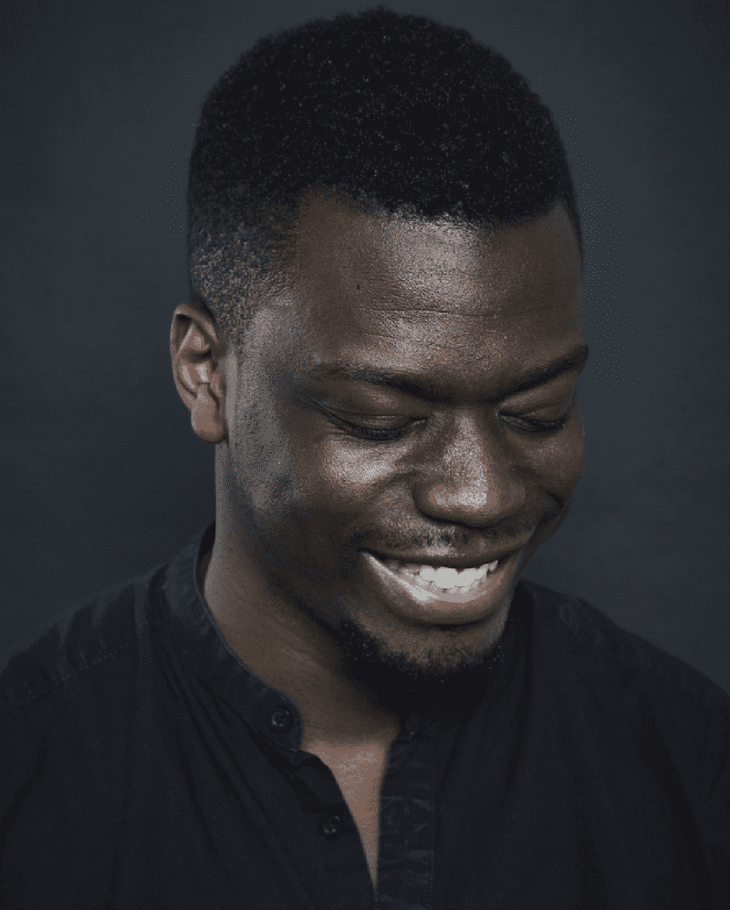 Ryan Calais Cameron Headshot. A black man in a dark shirt smiling and looking down against a dark grey background.