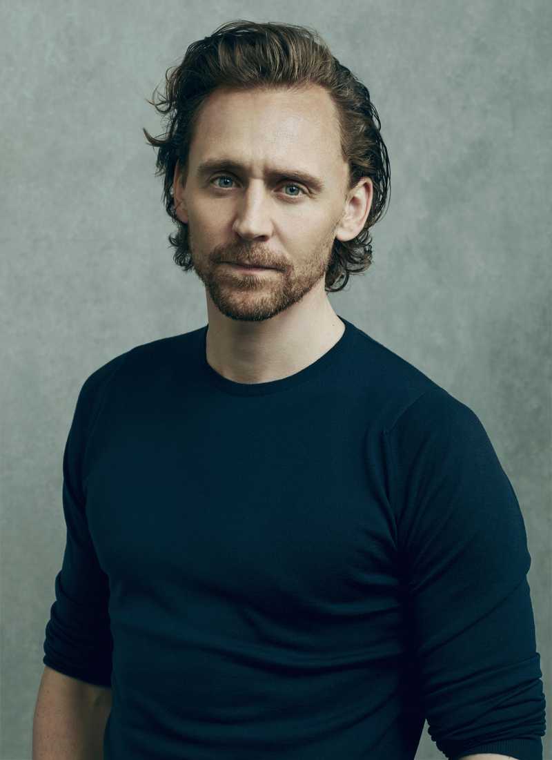 Tom Hiddleston Headshot by Disney. A white man, with dark brown hair and full goatee, standing against a pale sponged green background, wearing a deep blue top. 