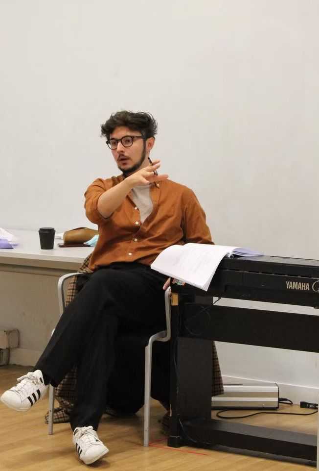 Rehearsal shot of Dewi Johnson, sat with script in hand, gesturing to actors out of the frame