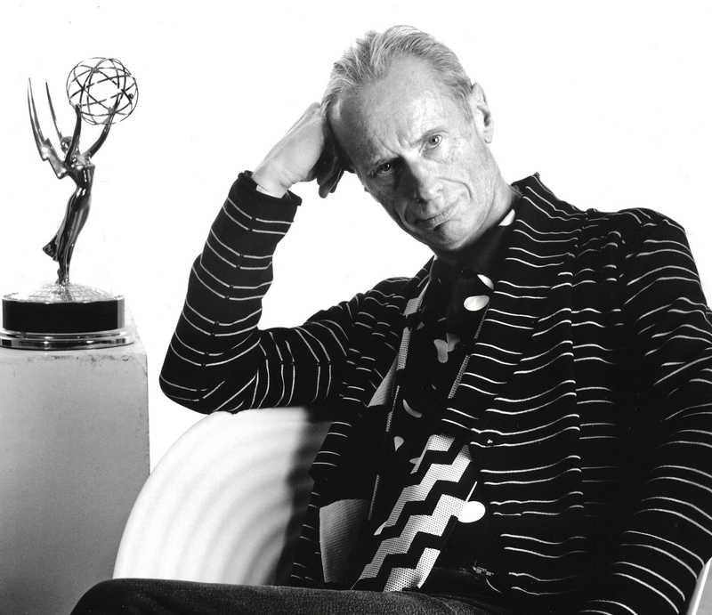 Image of Hugh Durrant dressed in a mix of stripes, with an Emmy award next to him. Black & White photo.